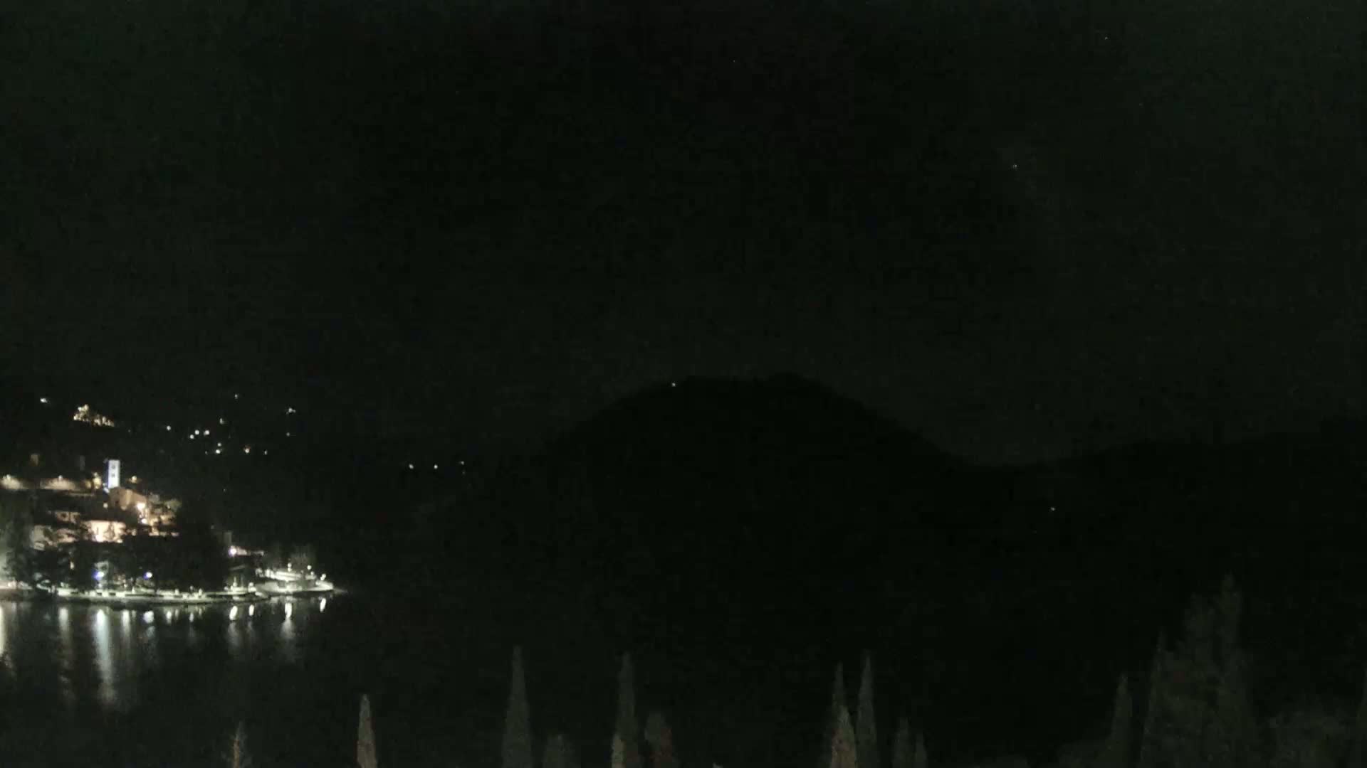 A dark nighttime scene shows a body of water reflecting scattered lights from a town nestled against a mountain.