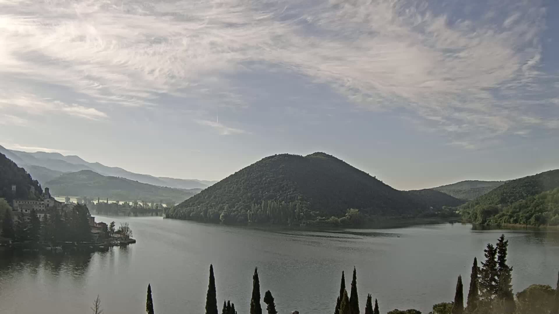 A calm lake is surrounded by mountains and hills under a partly cloudy sky.