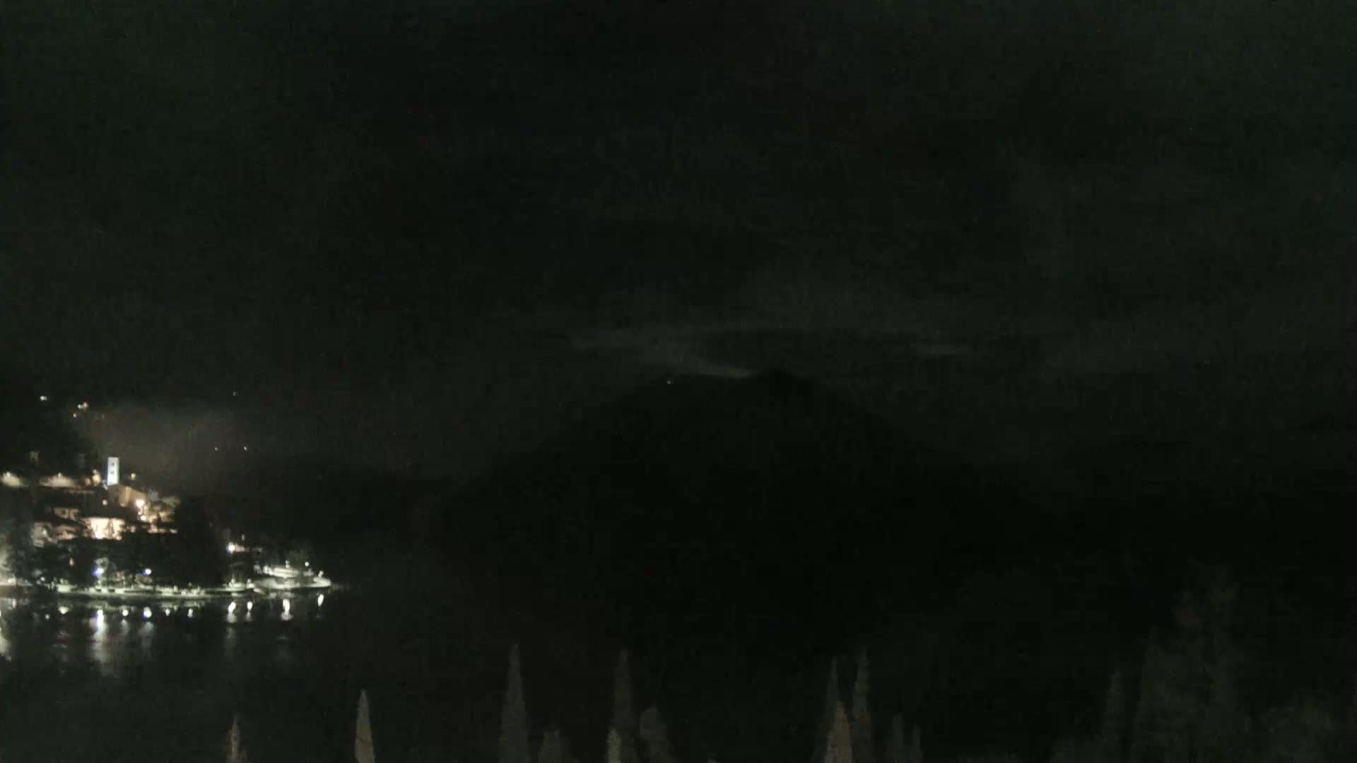 A nighttime view of a dark, calm body of water with a small town illuminated on the opposite shore, and a dark mountain or hill in the background under a cloudy sky.