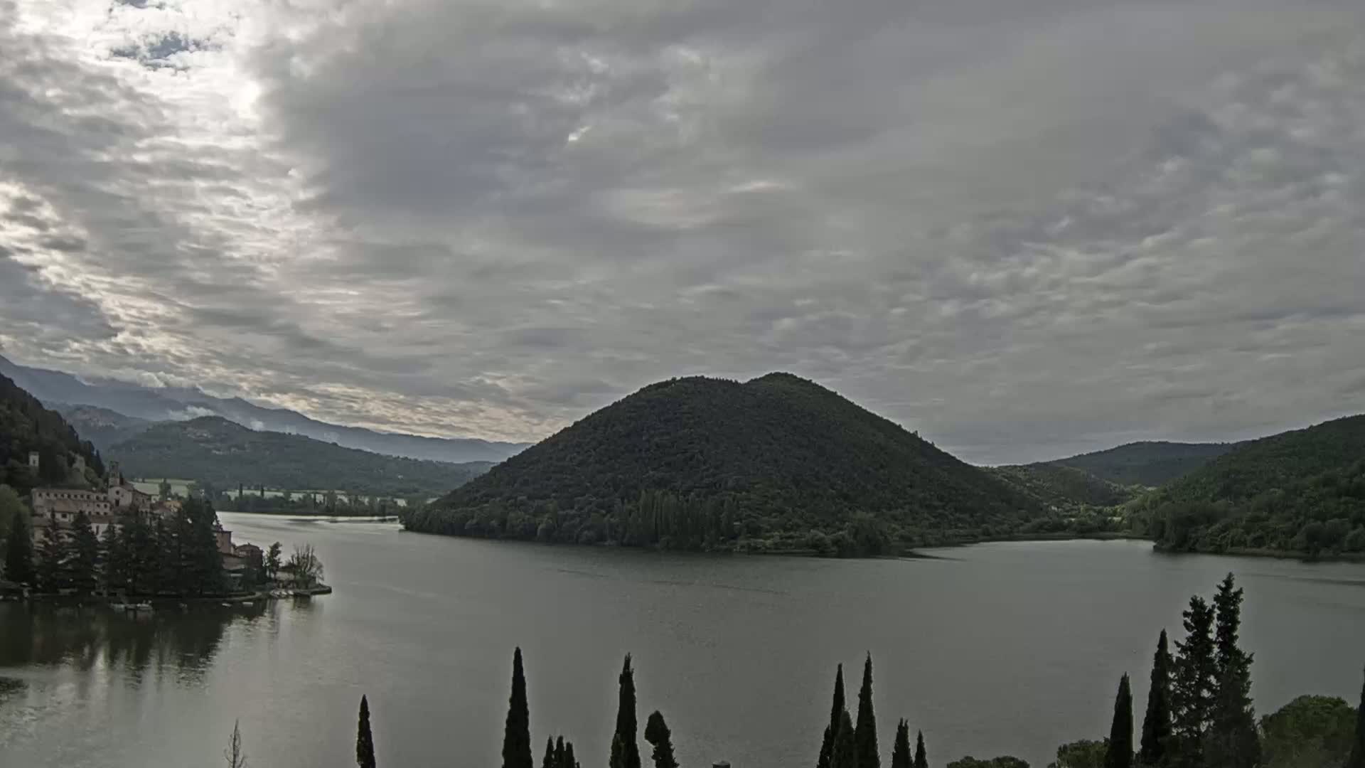A lake is seen, under a cloudy sky, with a large, dark hill in the center and a building complex on the left shore.