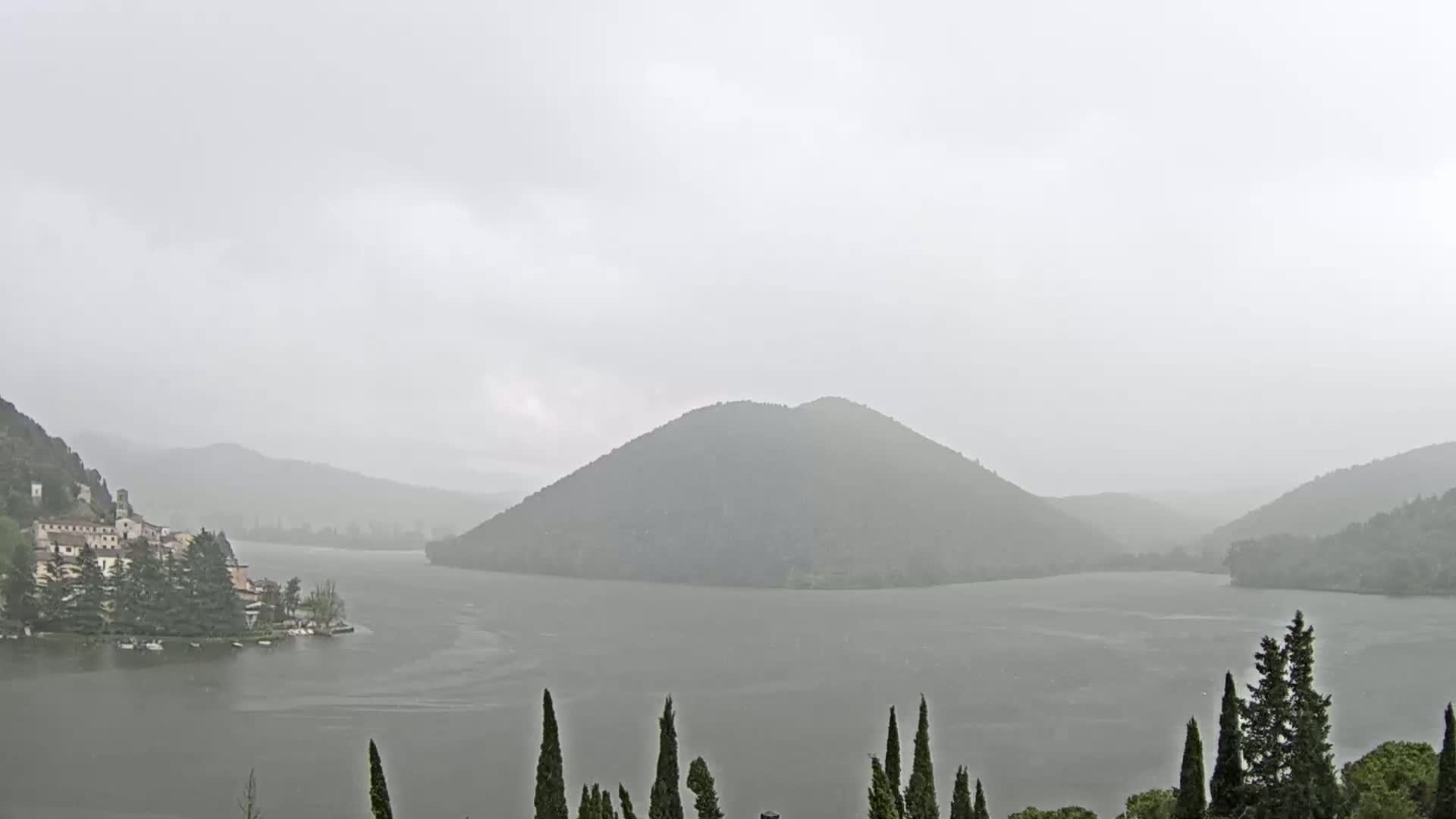 A lake is seen in the rain, surrounded by hills and a small village on the left shore, with dark evergreen trees in the foreground.