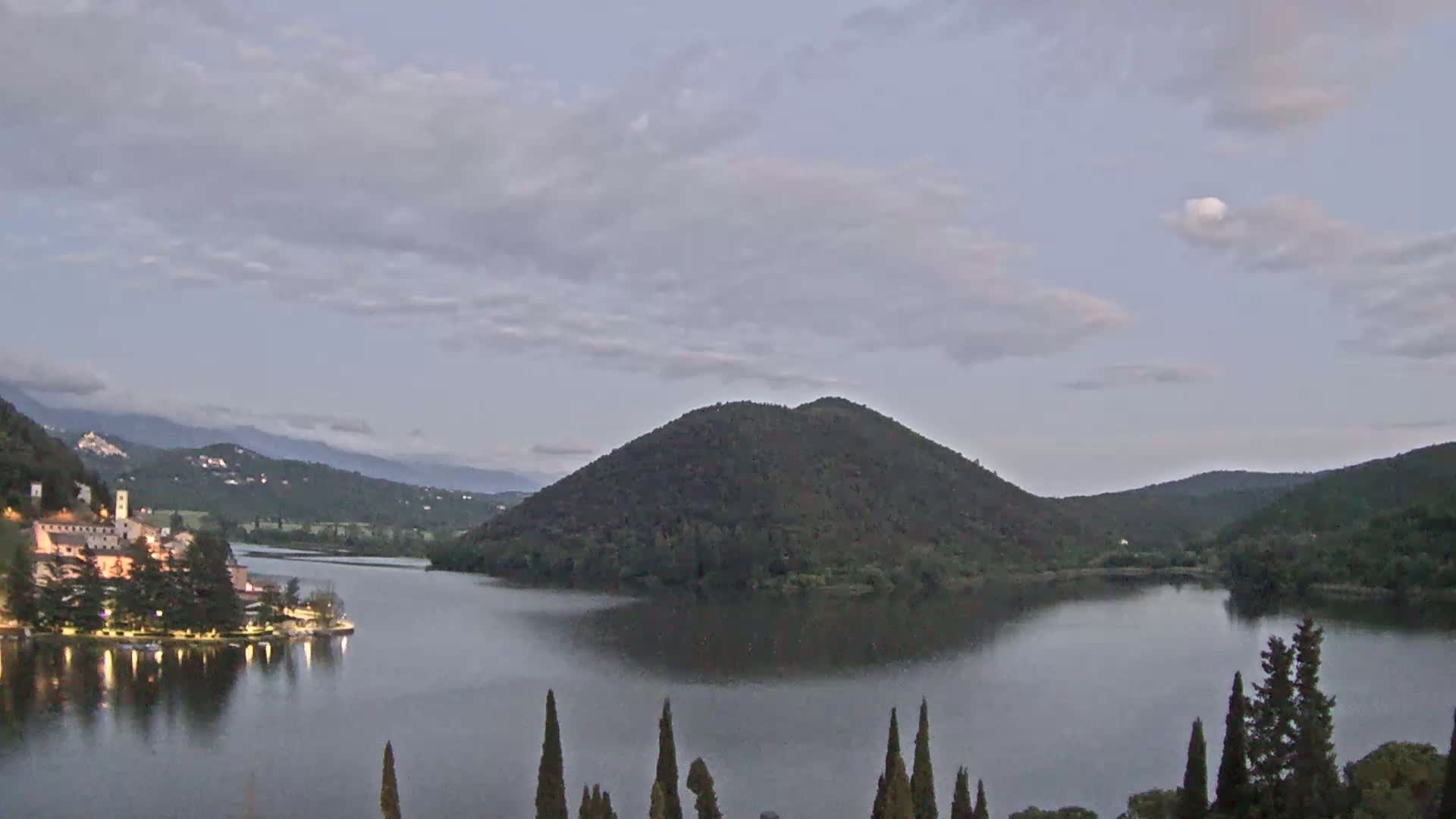 A calm lake at dusk is nestled between rolling hills under a partly cloudy sky.