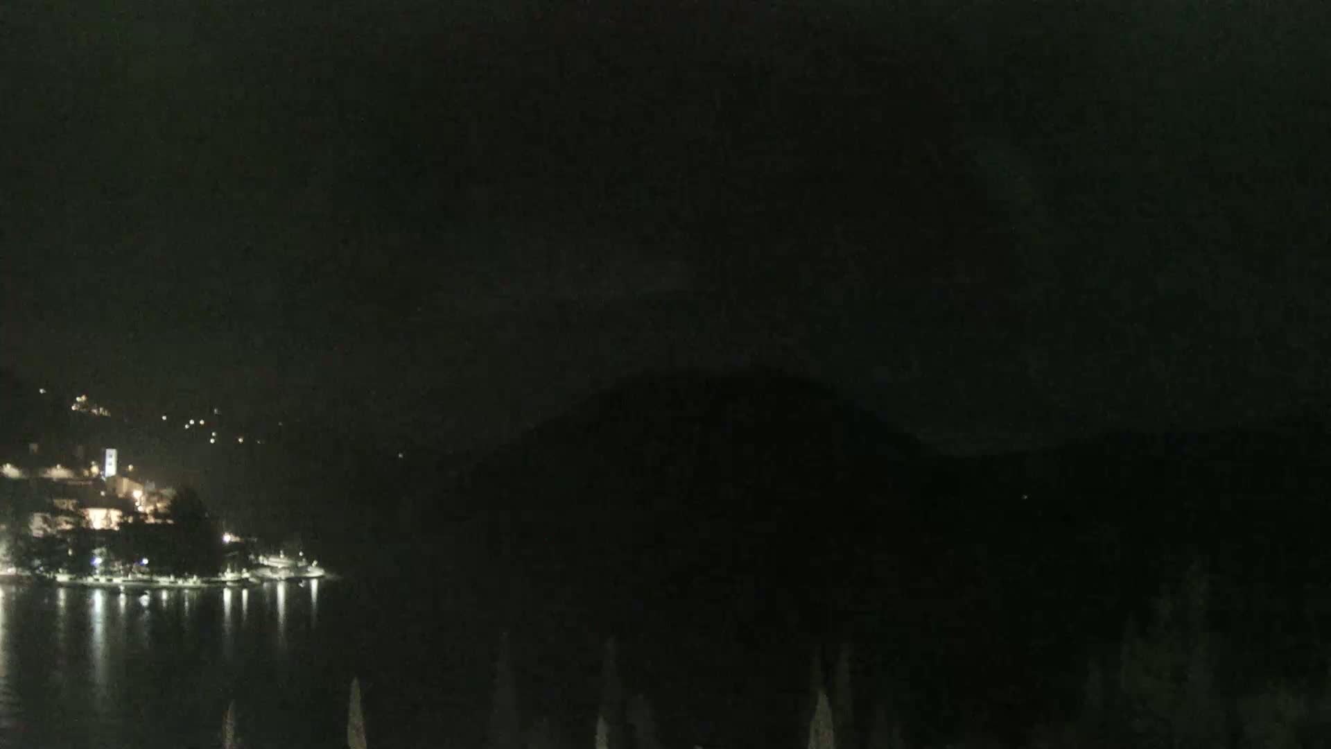 A dark, nighttime view shows a body of water reflecting the lights of a town nestled on a hillside, with dark mountains in the background under a mostly clear sky.
