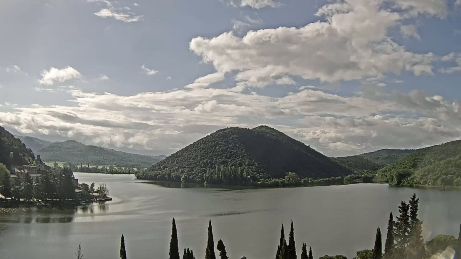 A lake nestled in a valley between green hills under a partly cloudy sky is viewed from a distance.