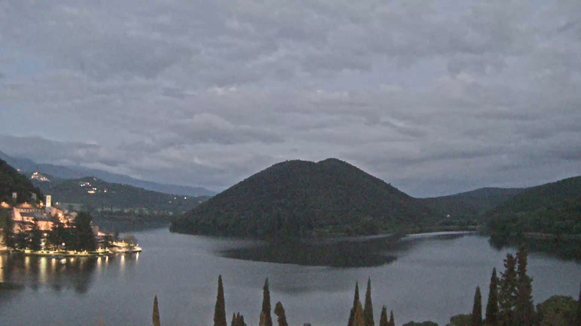 A tranquil lake at dusk is nestled between rolling hills under a cloudy sky, with a small village and island visible on the shore.