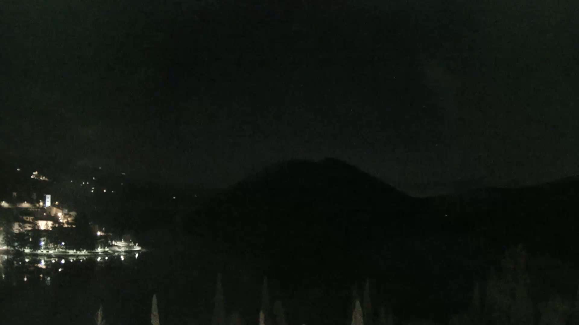 A nighttime view shows a dark, silhouetted mountain overlooking a body of water with a small, lit town on the opposite shore.