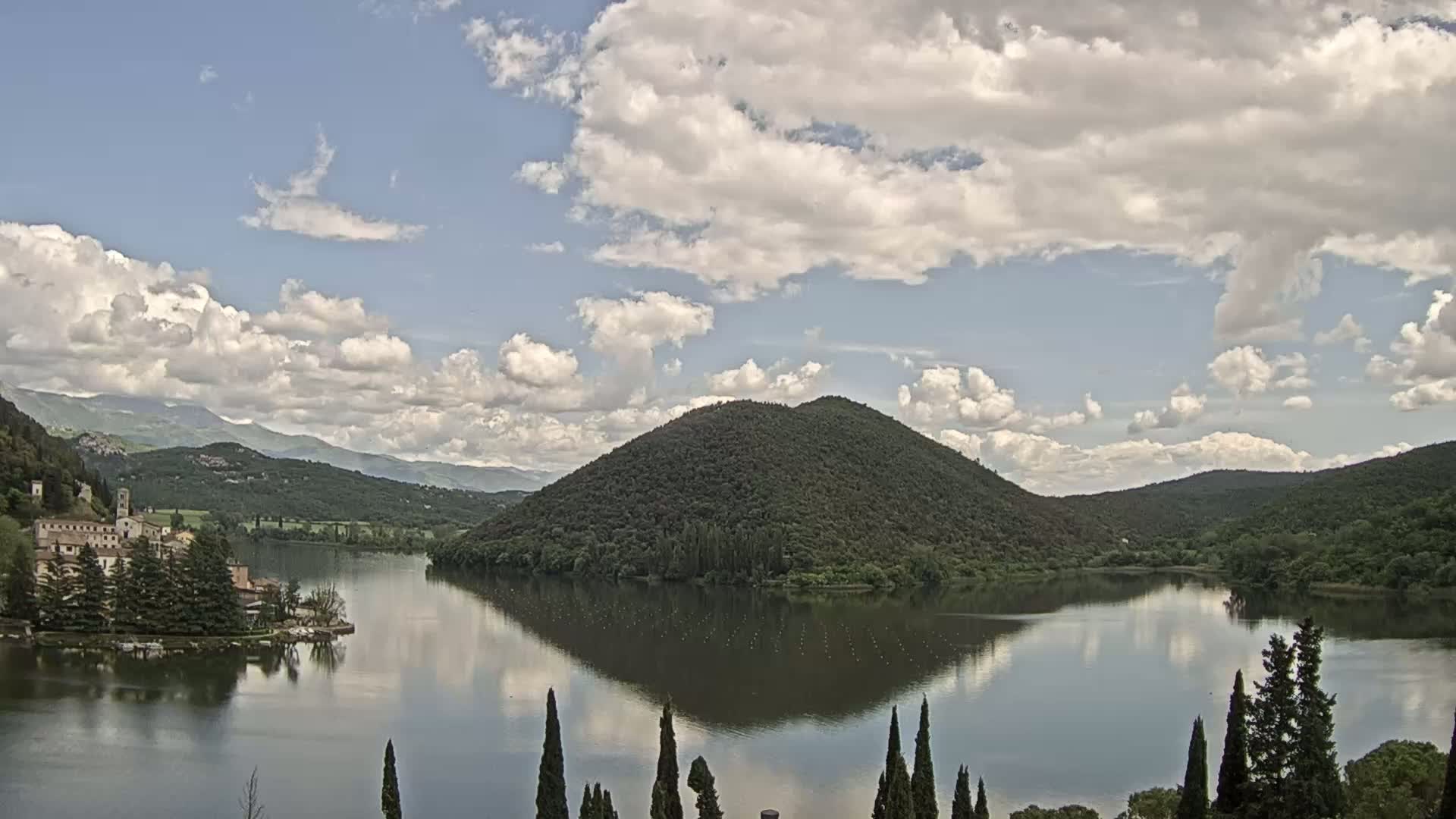 A calm lake reflects a tree-covered hill and distant mountains under a partly cloudy sky.