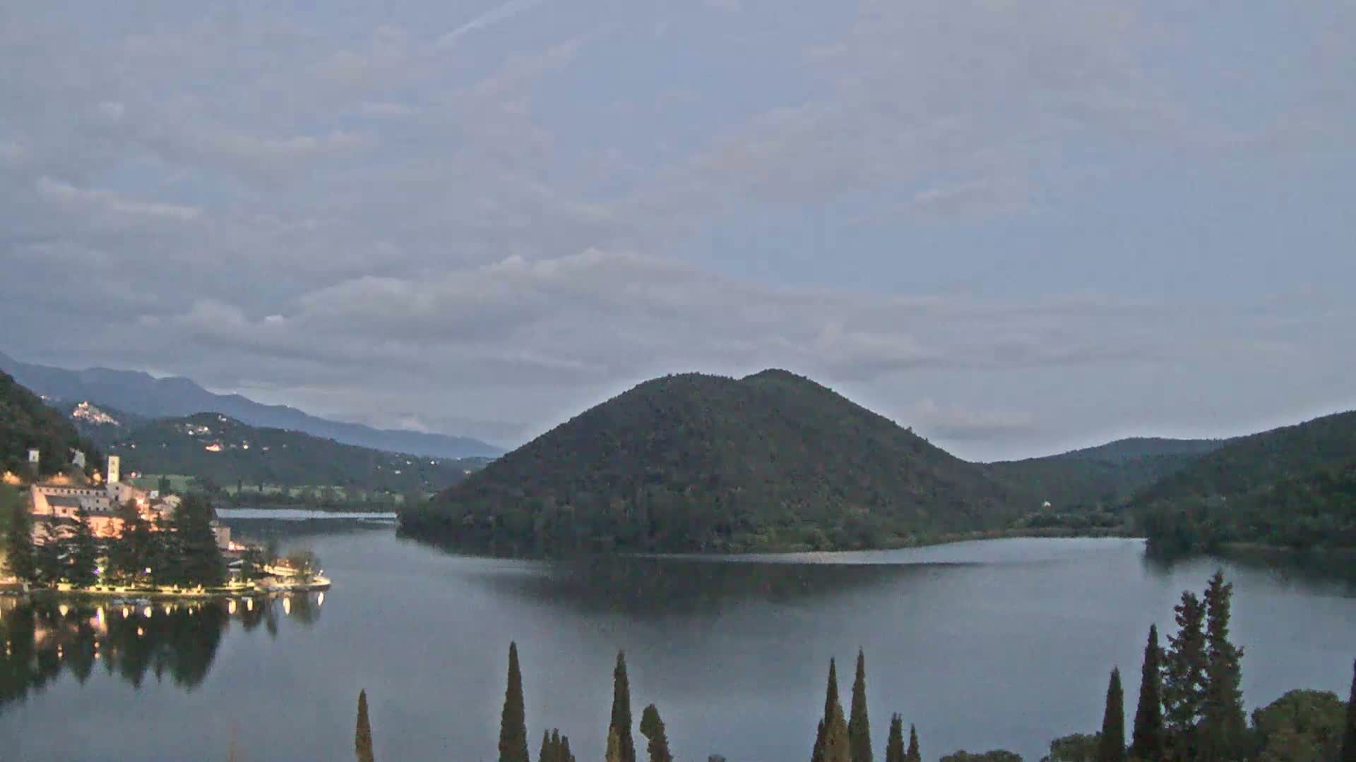 A lake at dusk, under a cloudy sky, is surrounded by hills and a village with lights visible along its shore.