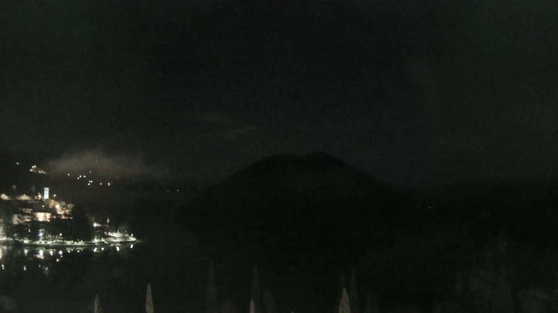 A nighttime view of a lake, with a town and a mountain silhouetted against a dark, cloudy sky.