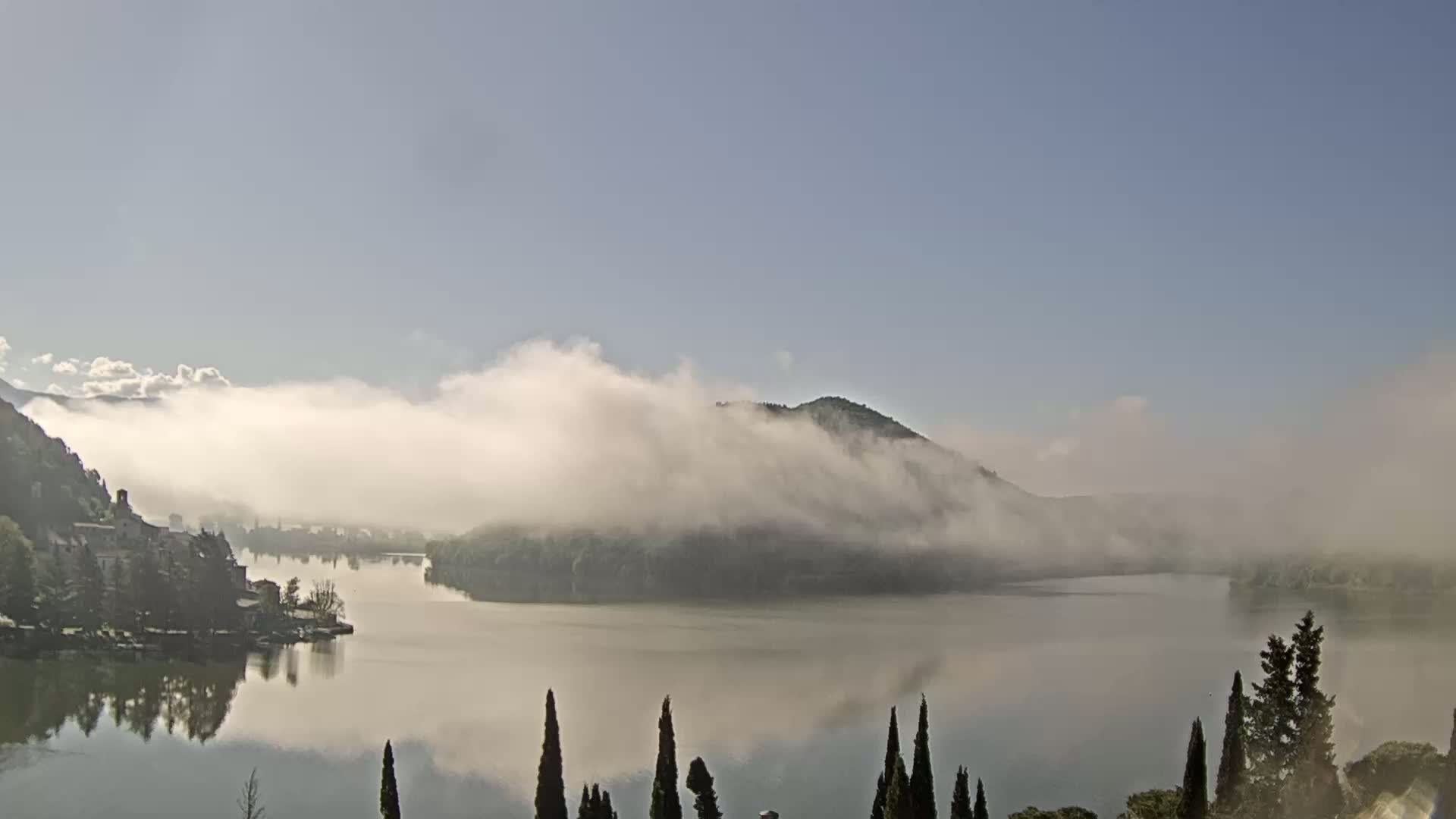 A calm lake is shrouded in morning fog, with hills and a small village visible in the background.