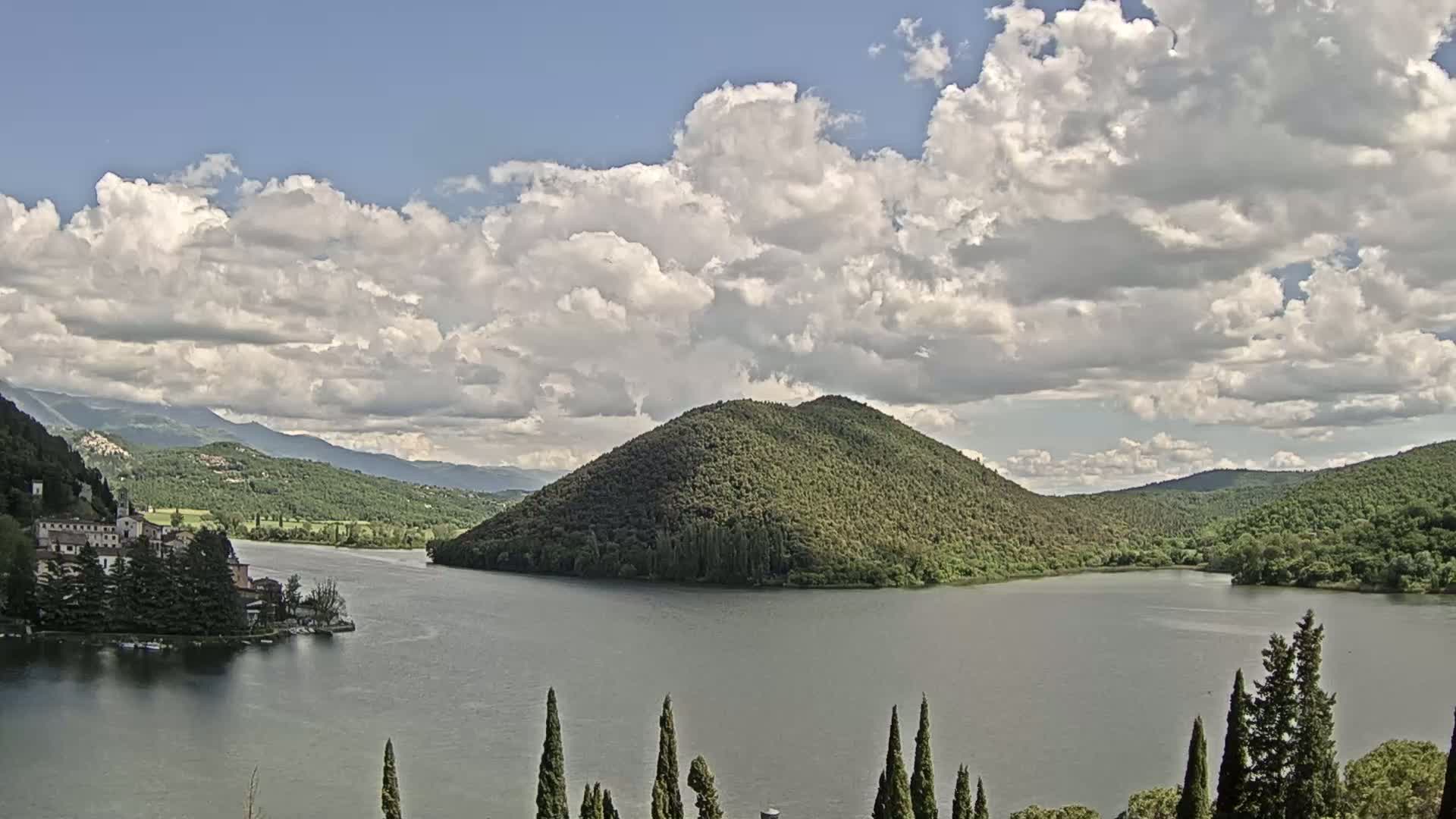A lake surrounded by green hills and a building complex on the lakeshore, under a partly cloudy sky.