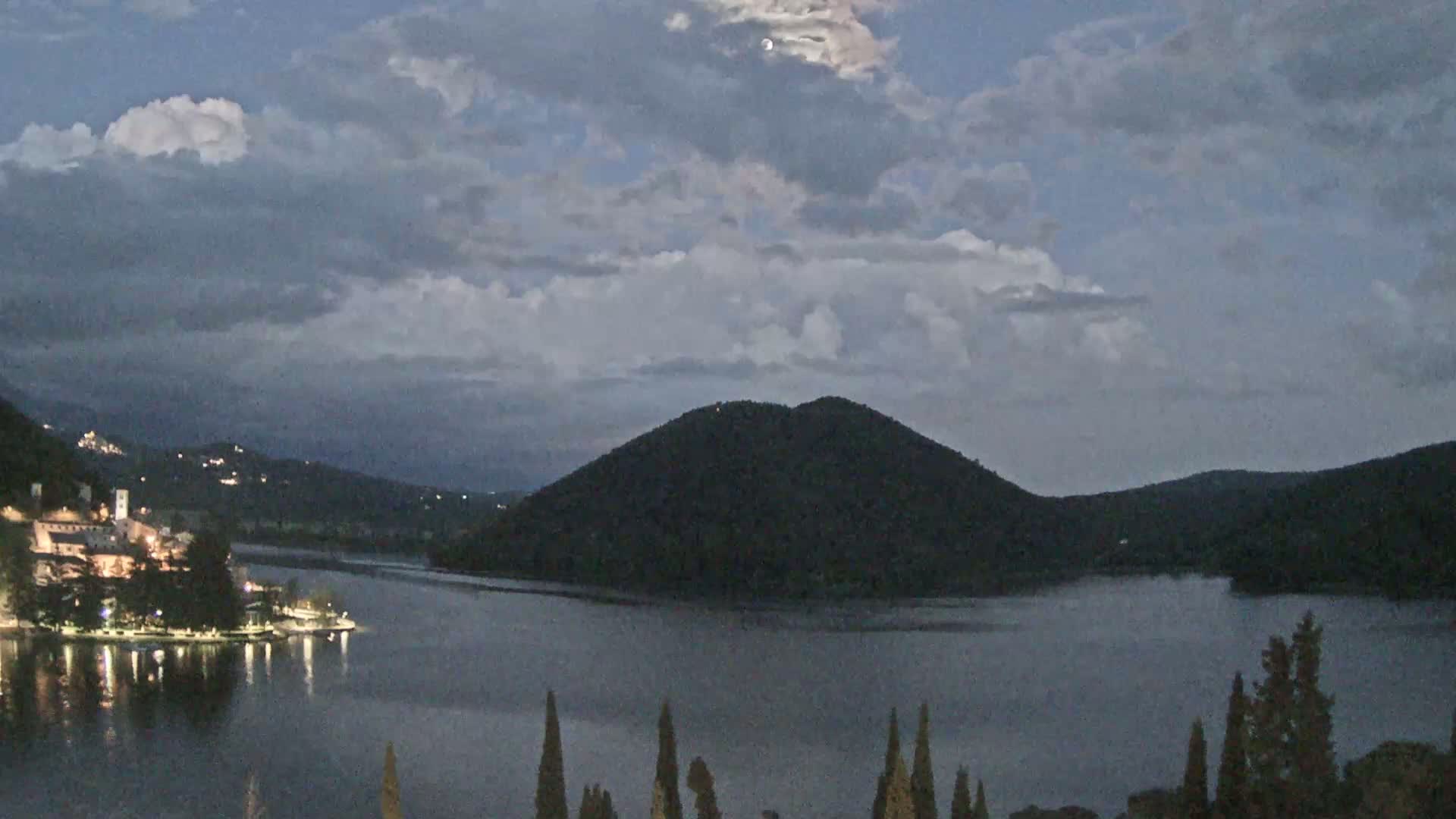 A moonlit lake at dusk, under a partly cloudy sky, is surrounded by dark hills and a village with lights on the shore.
