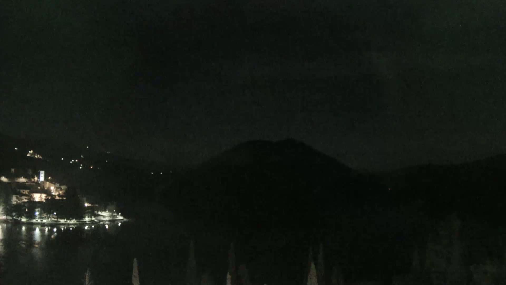 A dark, nighttime scene shows a body of water reflecting scattered lights from a town nestled between hills under a mostly cloudy sky.
