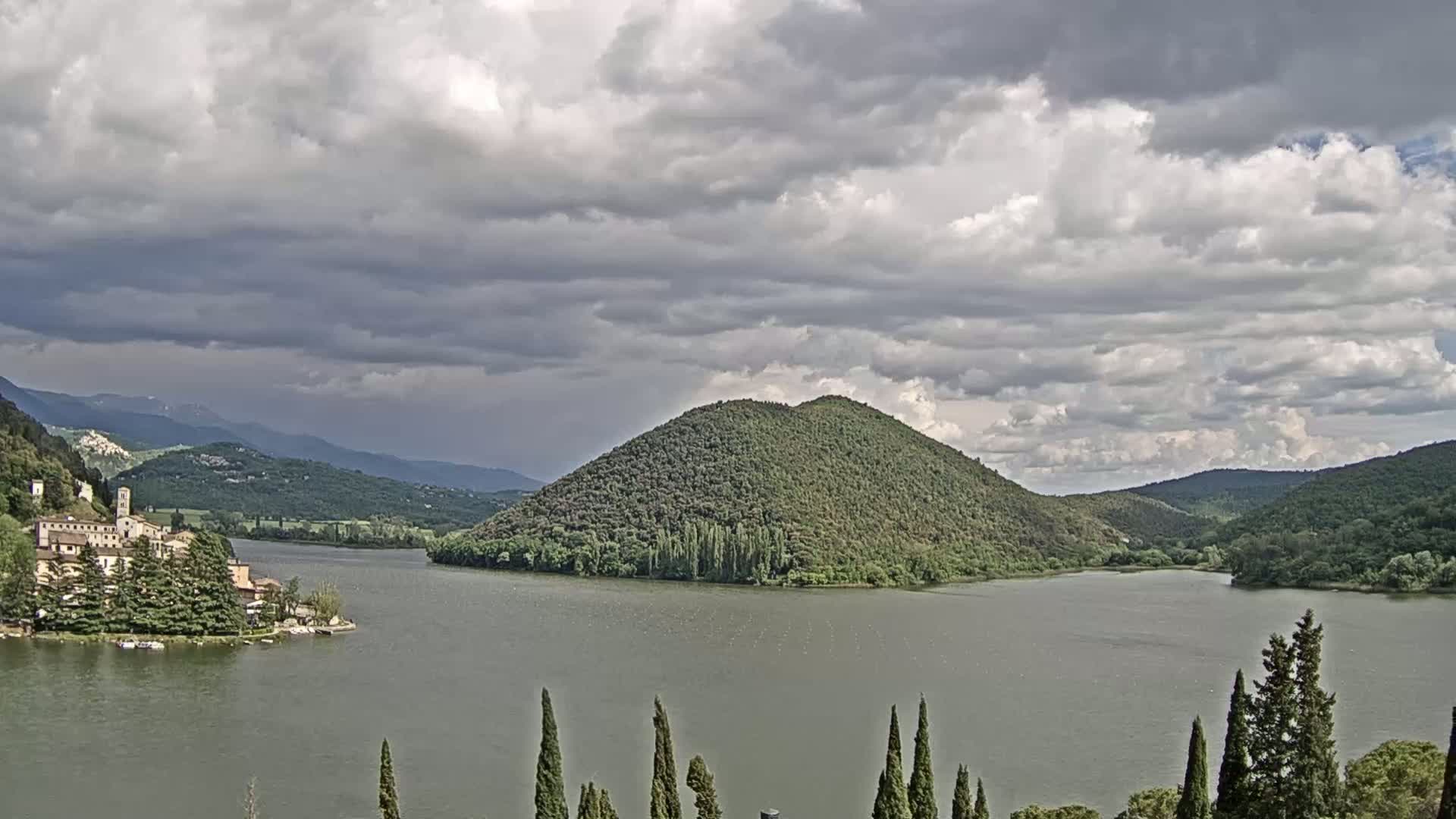 A lake is surrounded by green hills and a town under a cloudy sky.