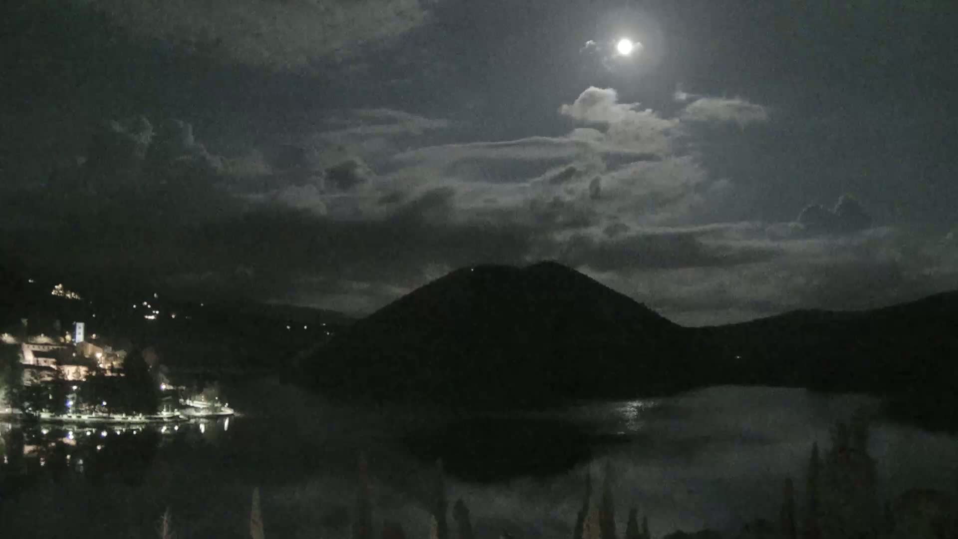 A moonlit night scene shows a calm lake reflecting the light, with a dark hill rising on the far side and a small town illuminated along the shore.