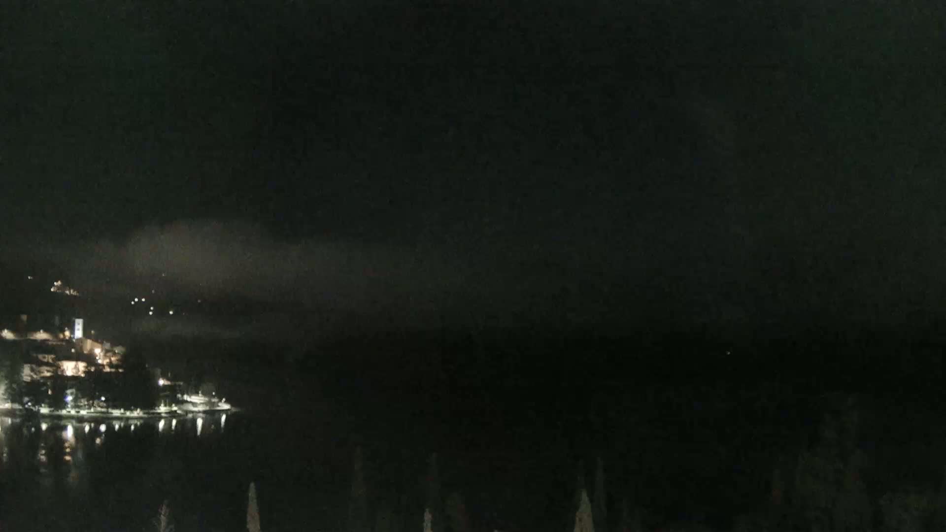 A nighttime view of a town's lights along a dark body of water under a cloudy, dark sky.