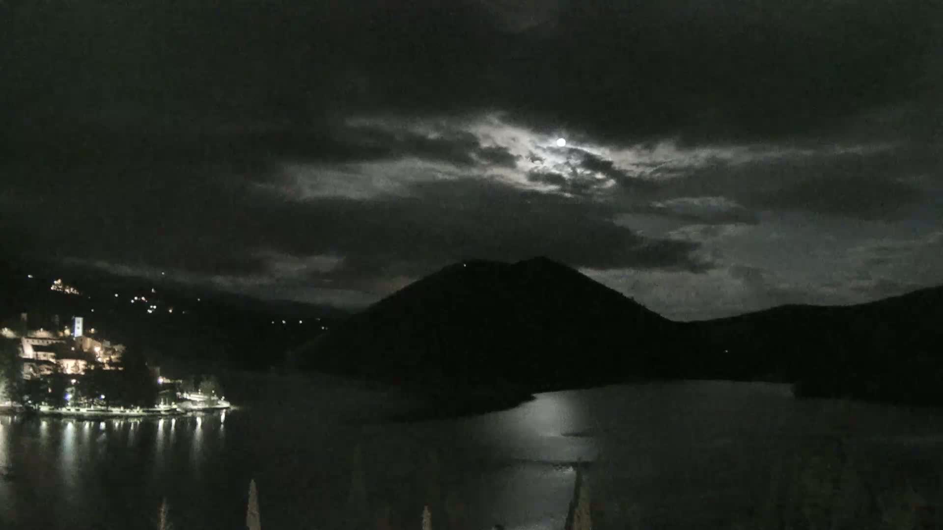 A moonlit night scene shows a body of water reflecting the light of a partially clouded moon, with a dark mountain in the background and a small, lit town on the shoreline.