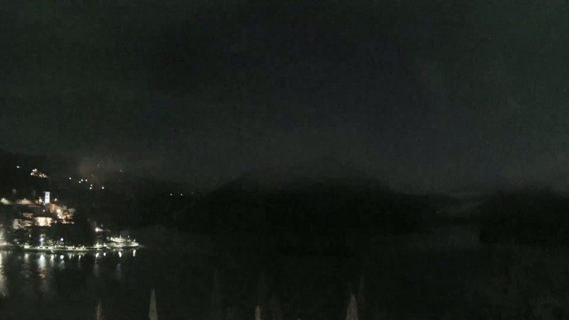 A nighttime view shows a dark body of water reflecting the lights of a town nestled in hills under a mostly clear, dark sky.