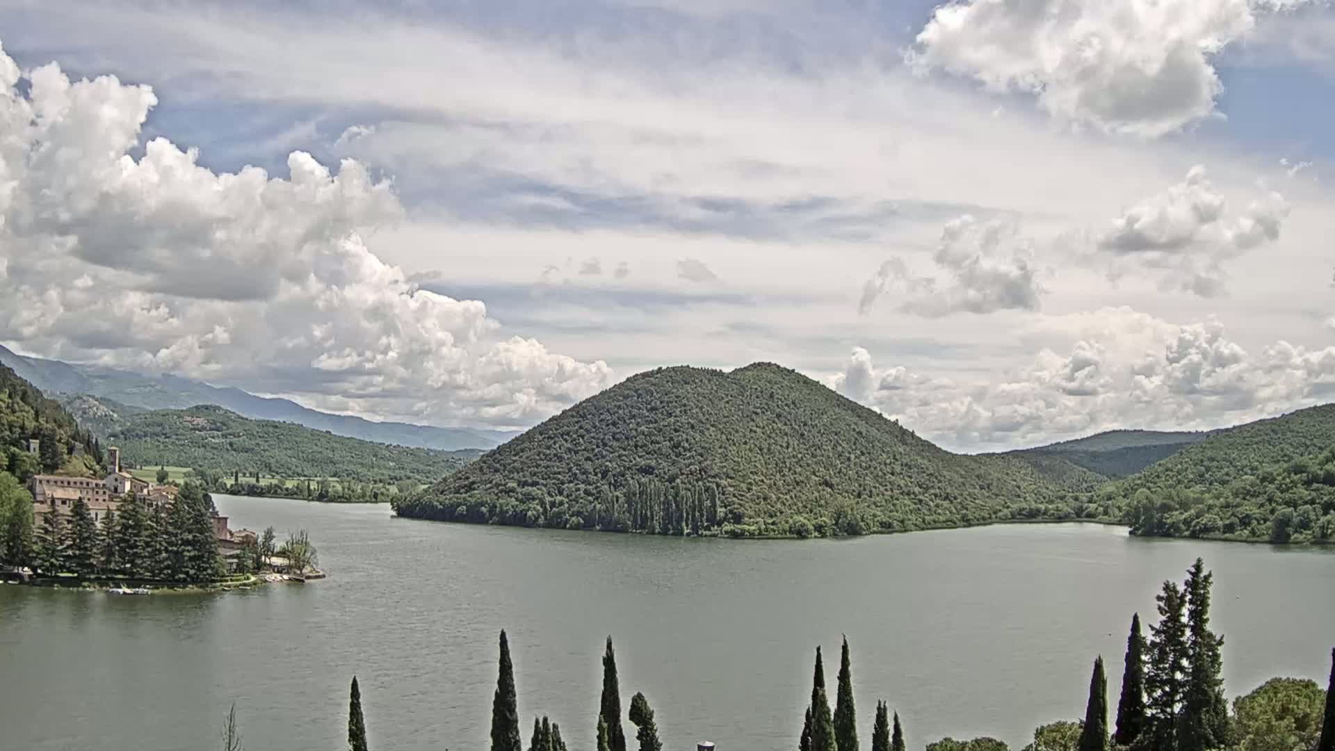 A lake surrounded by lush green hills and a building on an island under a partly cloudy sky.