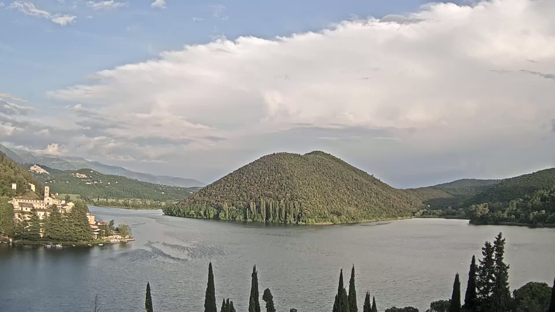 A lake with a tree-covered island is surrounded by hills and mountains under a partly cloudy sky.