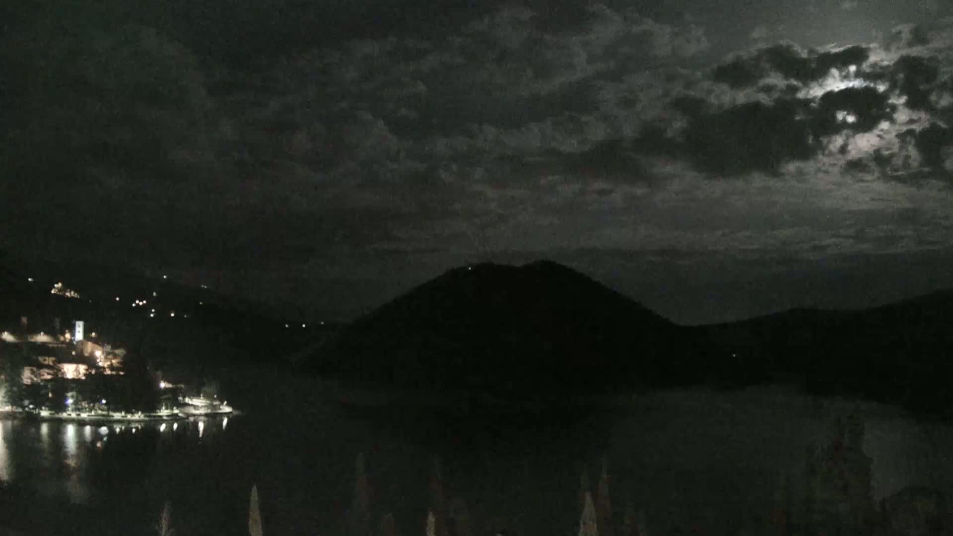 A dark, moonlit night scene shows a body of water reflecting the lights of a town nestled against a hill, with a larger, silhouetted hill rising beyond.