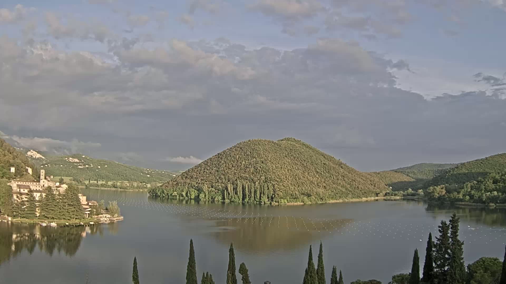 A lake, partially surrounded by green hills and a stone building, is visible under a partly cloudy sky.