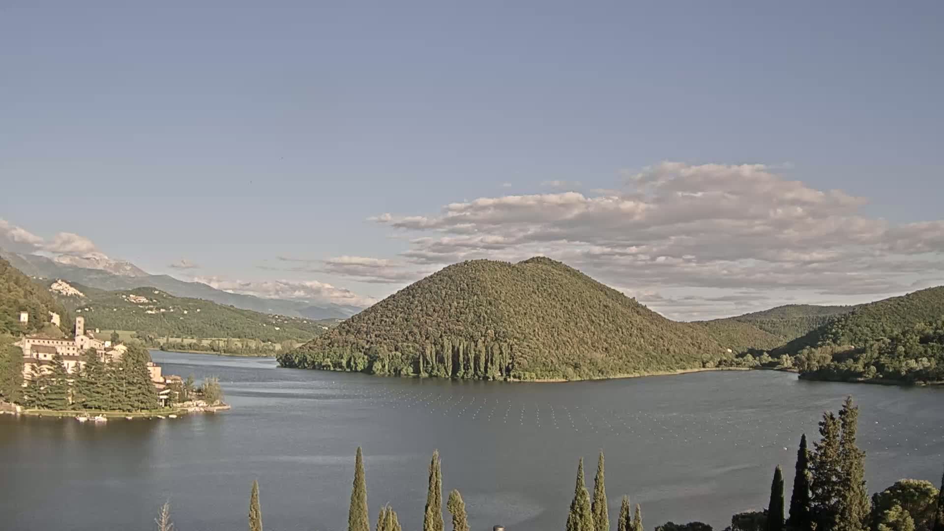 A calm lake is nestled among green hills and mountains under a partly cloudy sky.