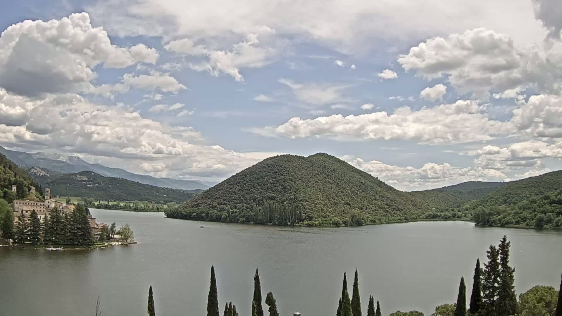 A lake is surrounded by green hills and mountains under a partly cloudy sky, with a building complex situated on the lakeshore to the left.