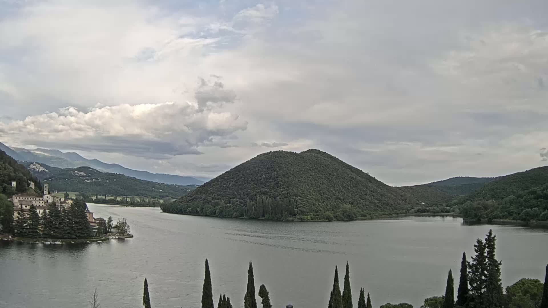 A lake surrounded by green hills and a building on the left shore under a mostly cloudy sky.