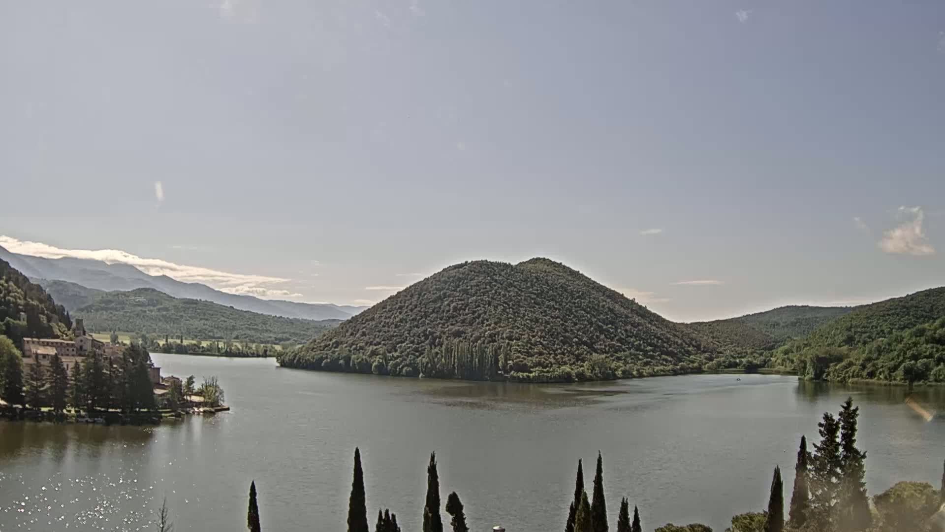A calm lake nestled between tree-covered hills and mountains under a mostly clear sky.