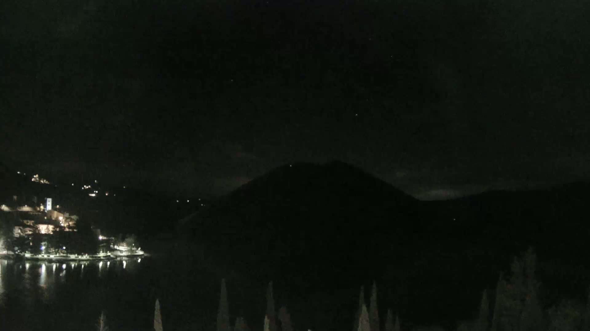 A nighttime view shows a dark body of water reflecting the lights of a small town nestled on a hillside, with a large, dark mountain in the background under a clear, dark sky.