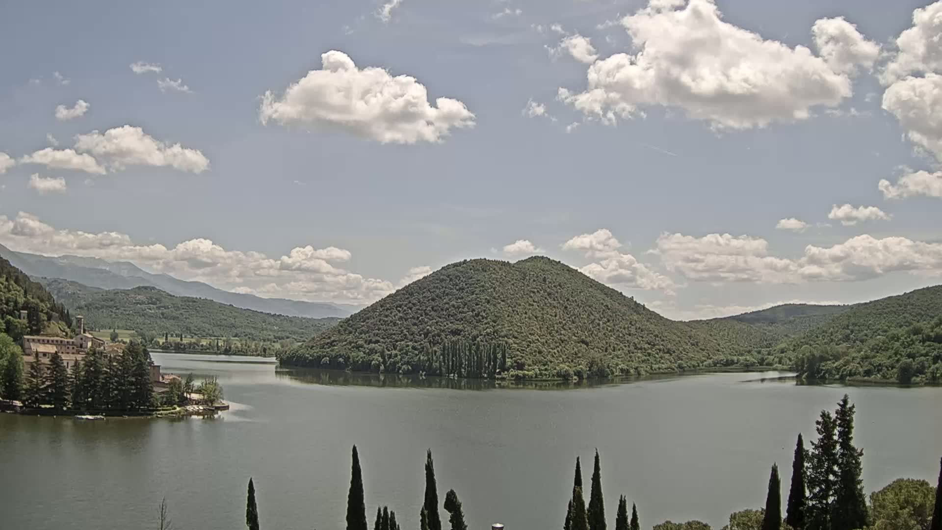 A calm lake is surrounded by lush green hills and a stone building near the water's edge under a partly cloudy sky.