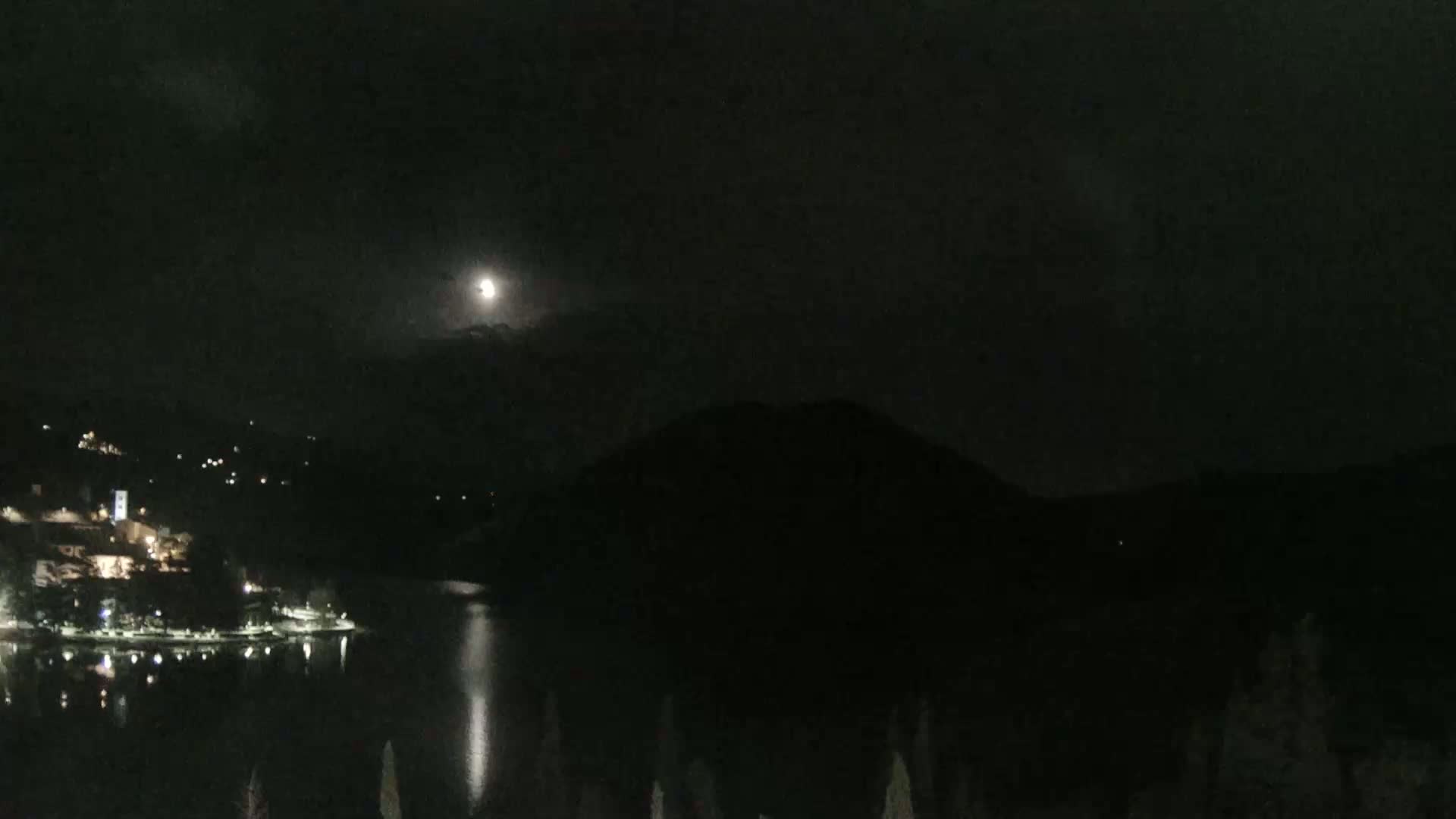 A bright moon illuminates a dark, calm lake reflecting lights from a town nestled against a mountain at night.