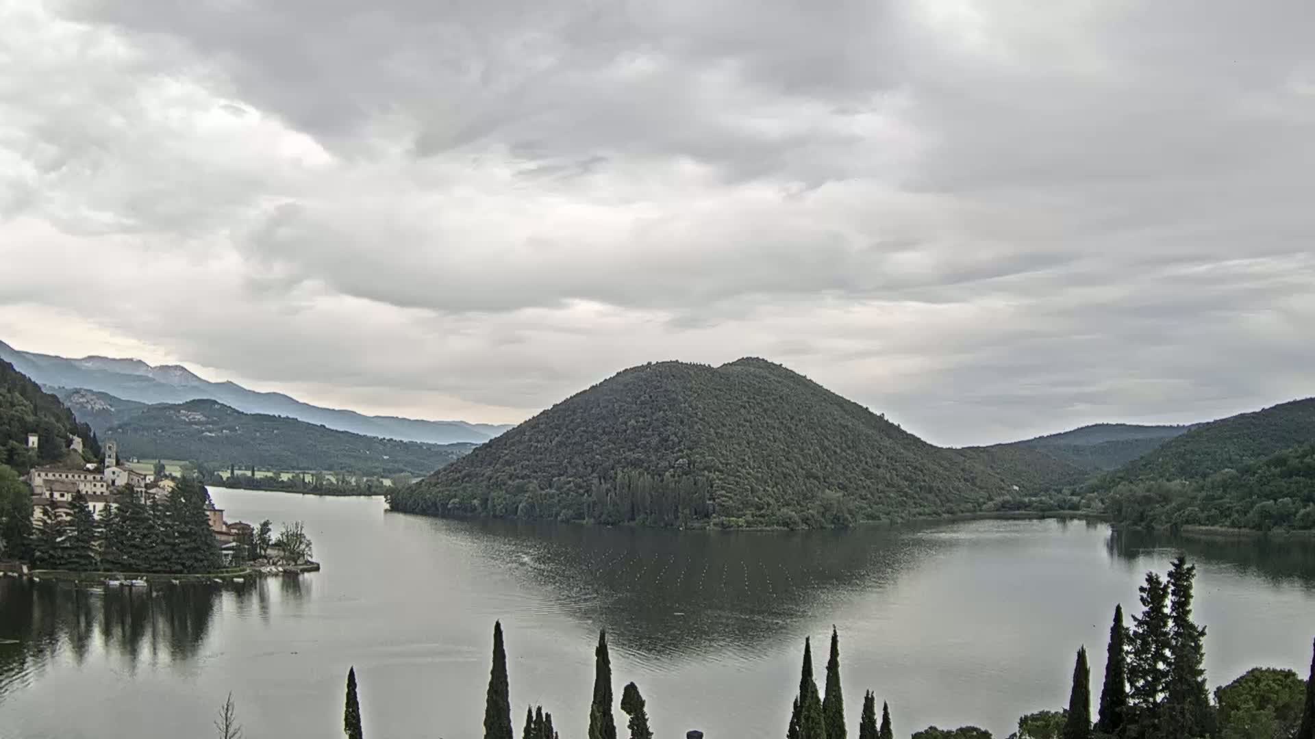 A calm lake is nestled between rolling hills under a cloudy sky, with a village situated on the lakeshore.