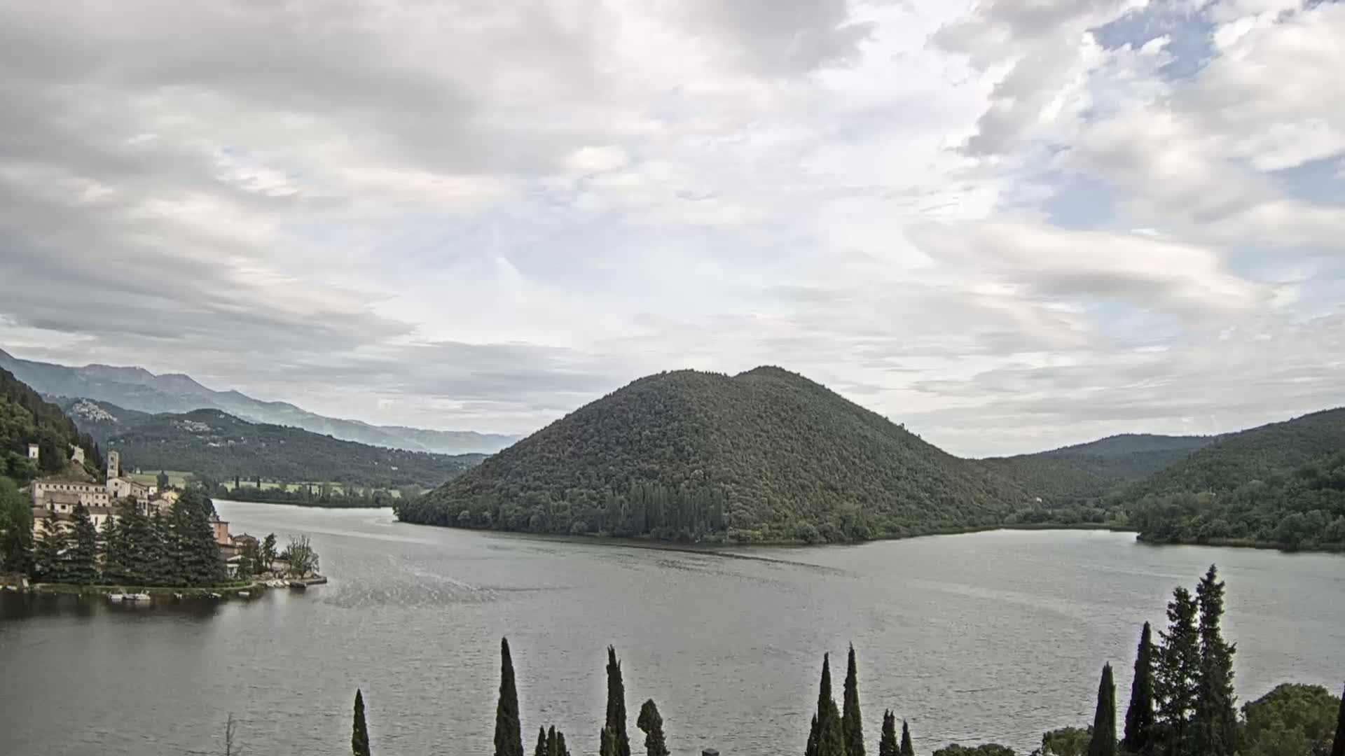 A lake is surrounded by lush green hills and mountains under a cloudy sky, with a village nestled on the lakeshore.