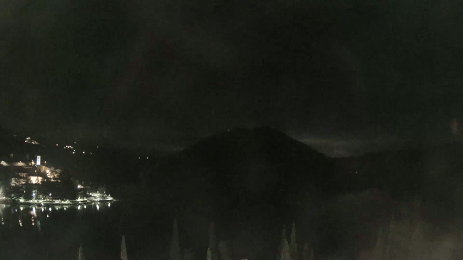 A nighttime view shows a dark mountain overlooking a calm lake with lights on the far shore, under a cloudy sky.