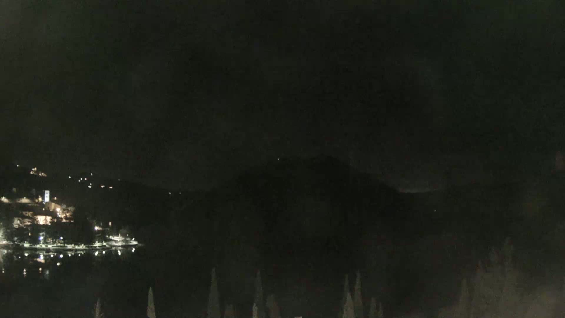 A nighttime view of a dark lake, with a hillside town faintly illuminated in the distance, under a mostly cloudy sky.