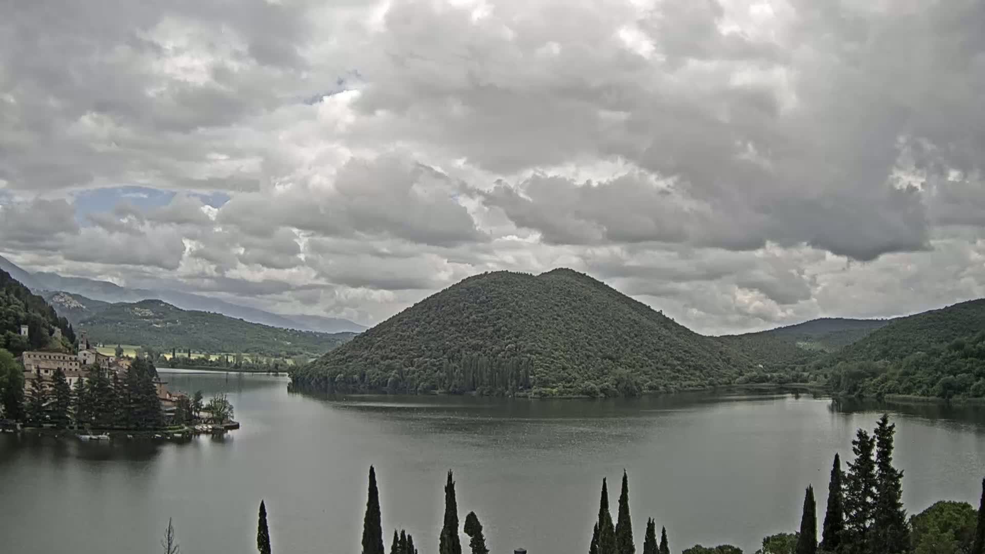 A lake, under a cloudy sky, is nestled between rolling hills, with a building visible on the lakeshore.