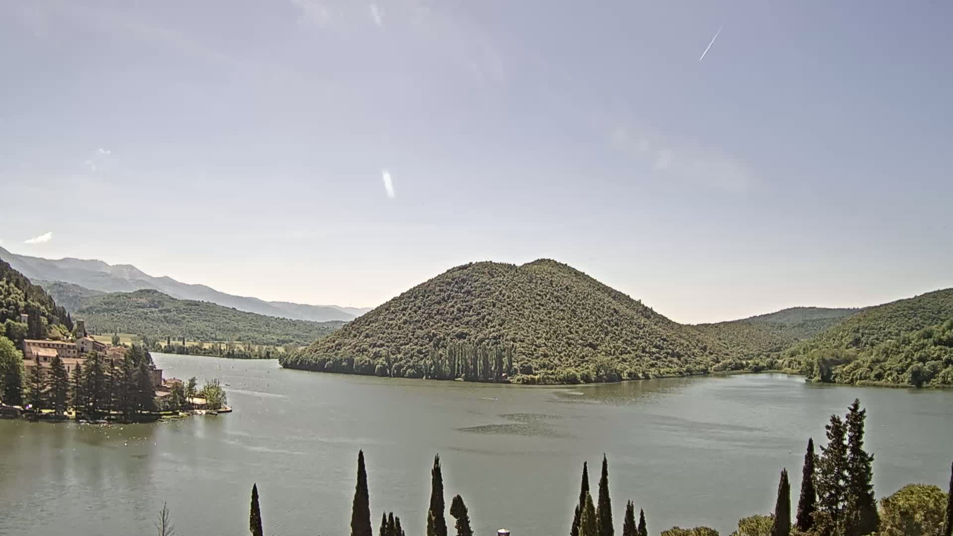 A lake surrounded by green hills and a building on the lakeshore under a mostly clear sky.