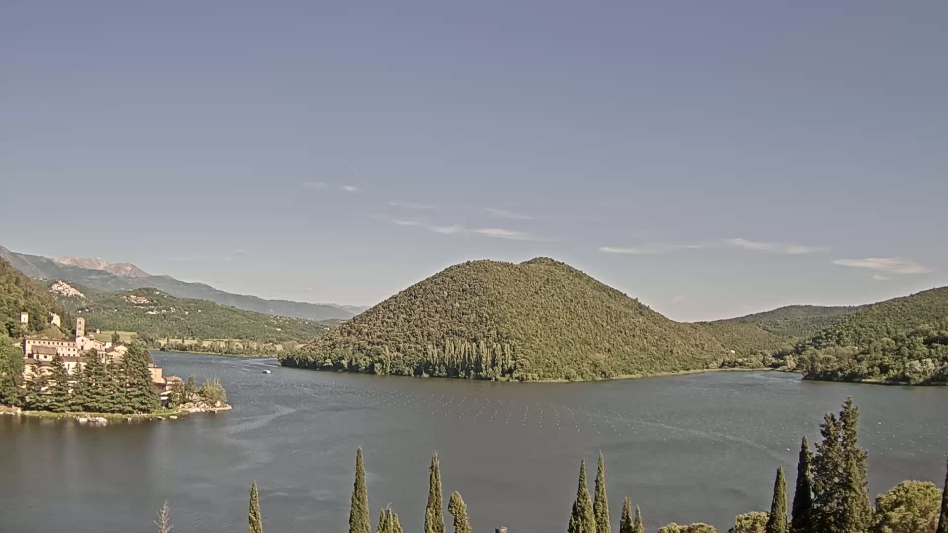A lake nestled in a valley between verdant hills and mountains under a clear, sunny sky, with a stone building situated on the lakeshore.