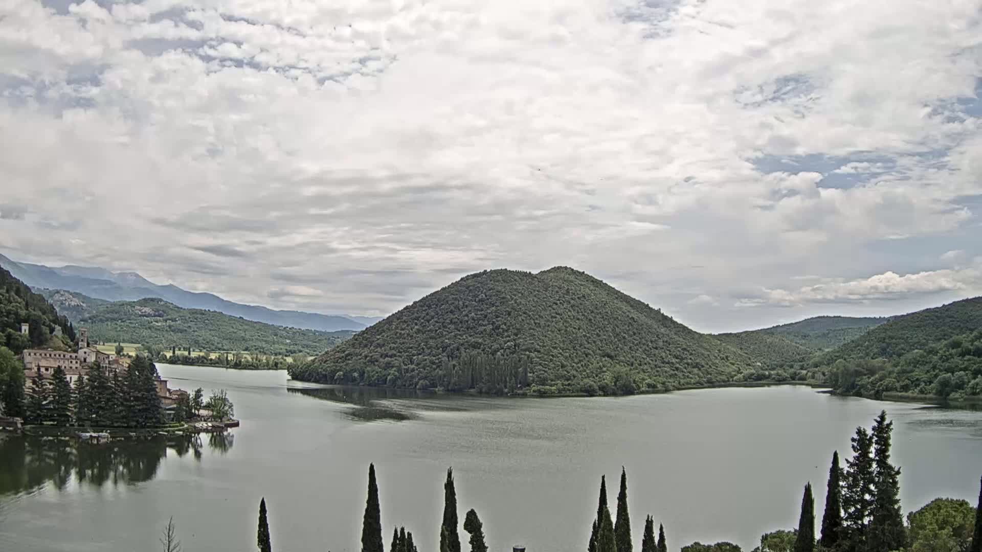 A lake surrounded by green hills and a stone building on the left, under a mostly cloudy sky.
