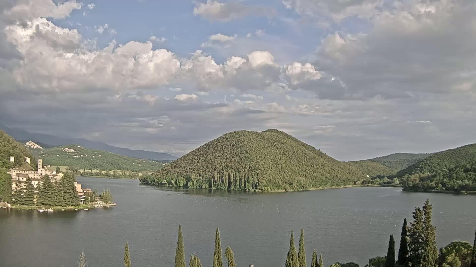 A lake surrounded by green hills and a building complex on the lakeshore under a partly cloudy sky.