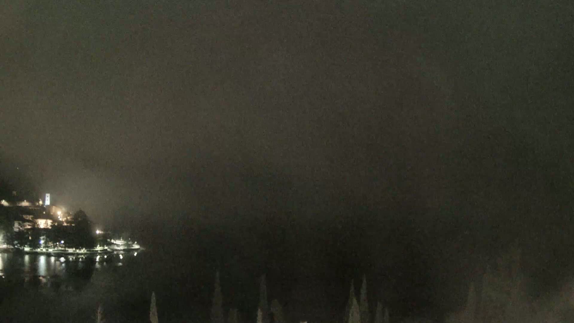 A nighttime view of a foggy, dark shoreline with a few lights visible on a hill overlooking the water.