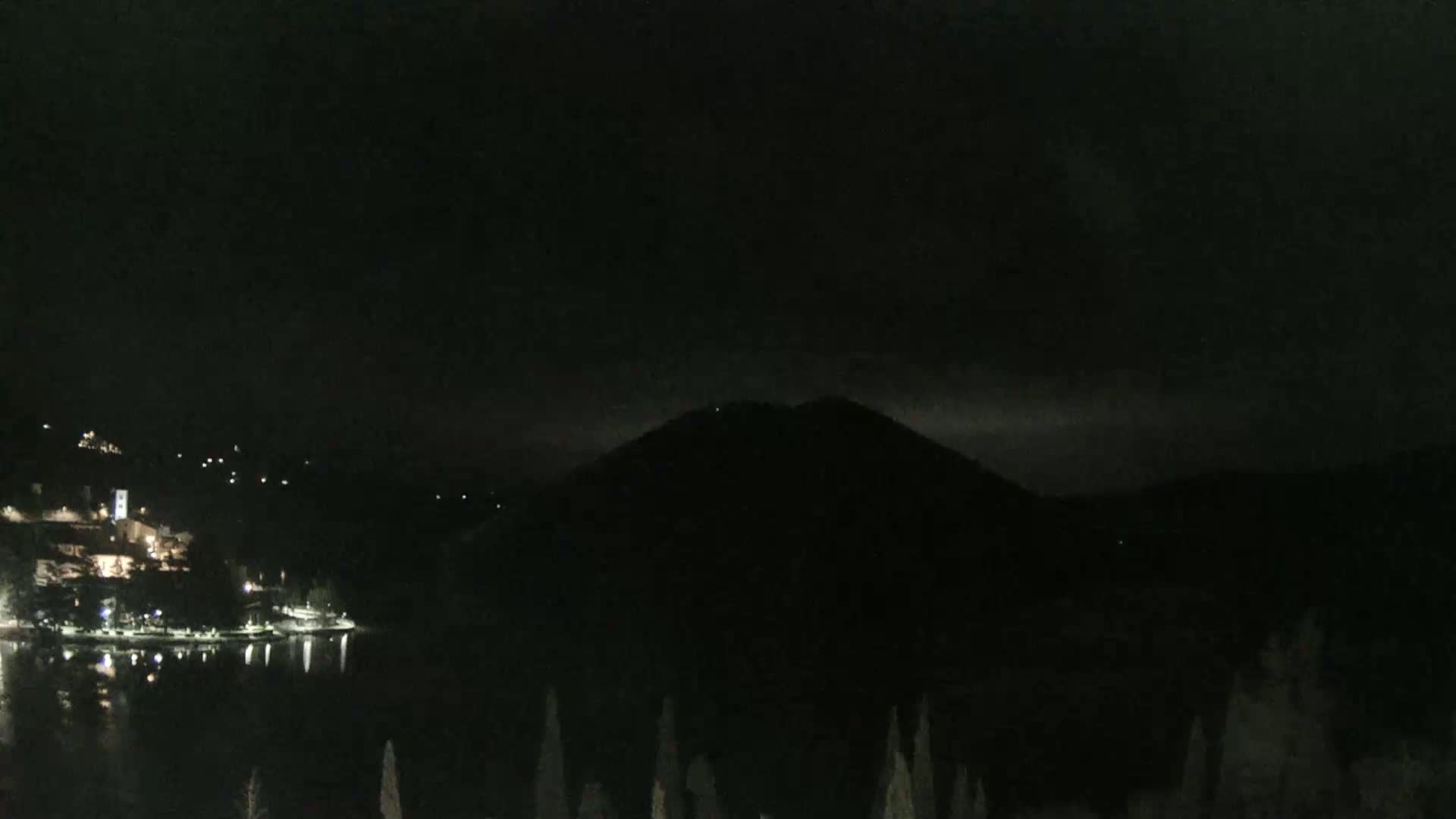 A dark, nighttime view shows a body of water reflecting the lights of a small town nestled against a large, dark hill under a mostly cloudy sky.