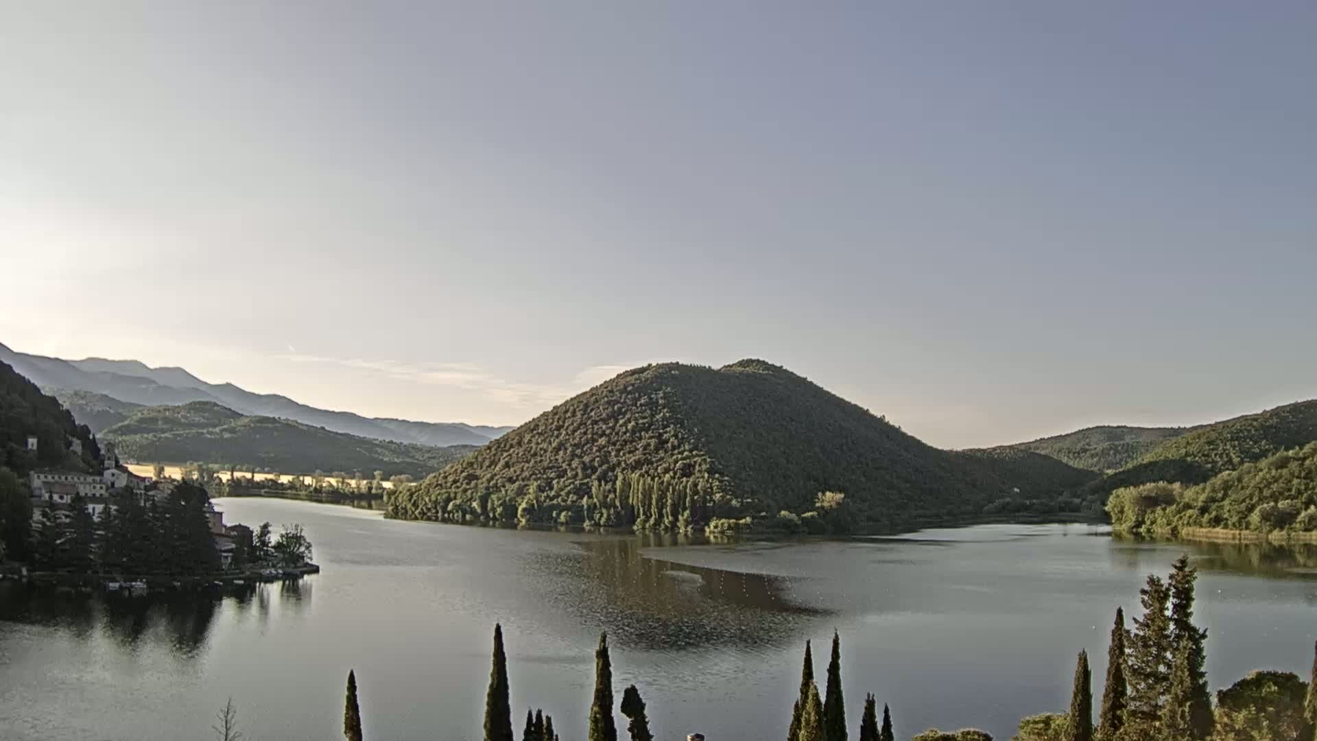 A calm lake is nestled in a valley between tree-covered hills under a clear, light sky.