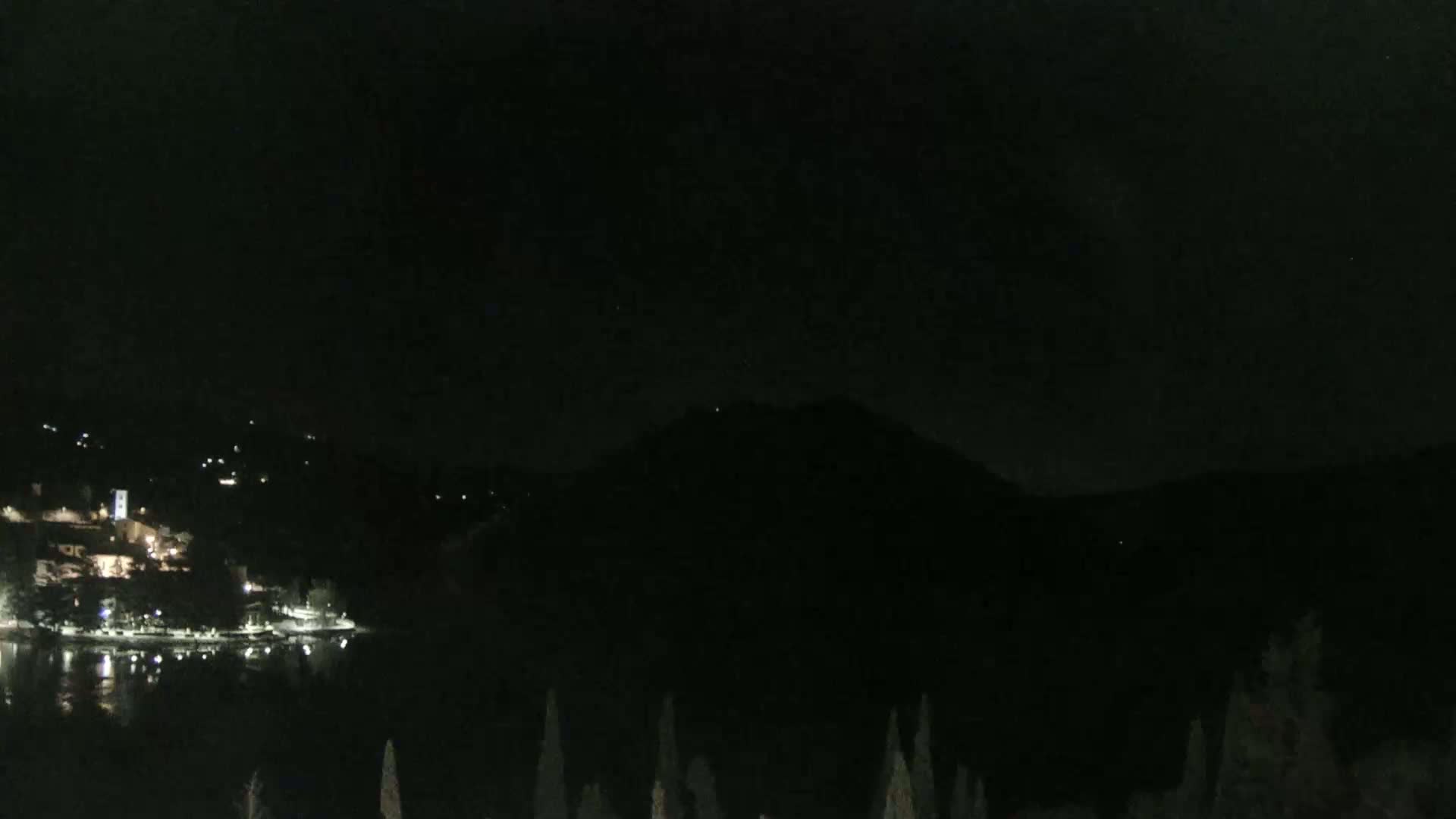 A nighttime view shows a dark lake reflecting the lights of a town nestled at the base of a large, dark mountain.