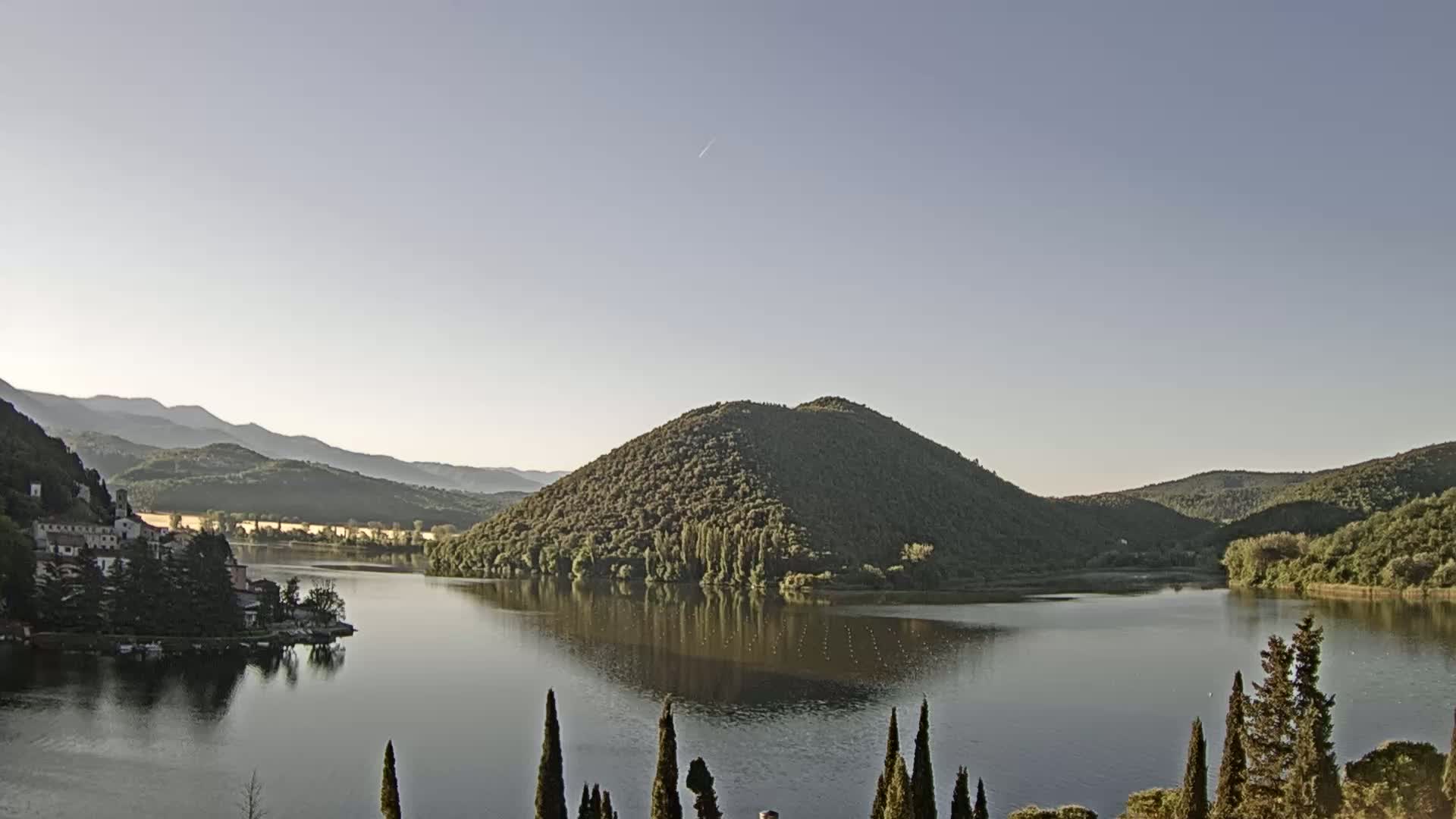 A calm lake reflects a tree-covered hill and distant mountains under a clear, light-blue sky.