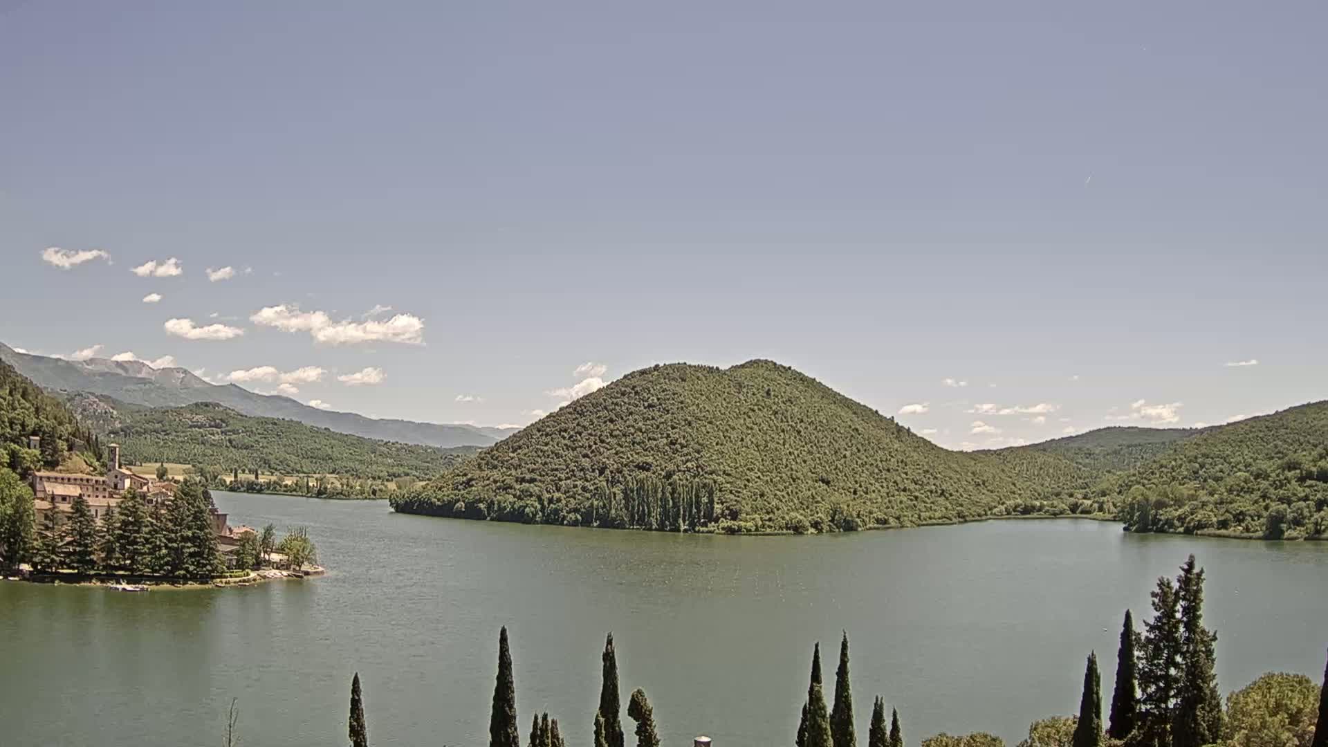 A calm lake is surrounded by green hills and mountains under a mostly clear sky with a few scattered clouds.