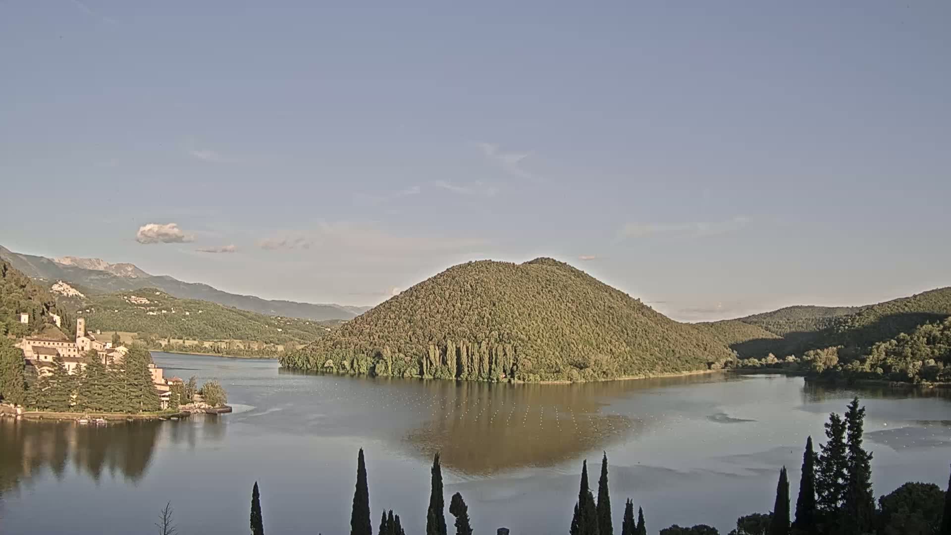 A calm lake is nestled between verdant hills under a clear, pale blue sky, with a building complex visible on the lakeshore to the left.