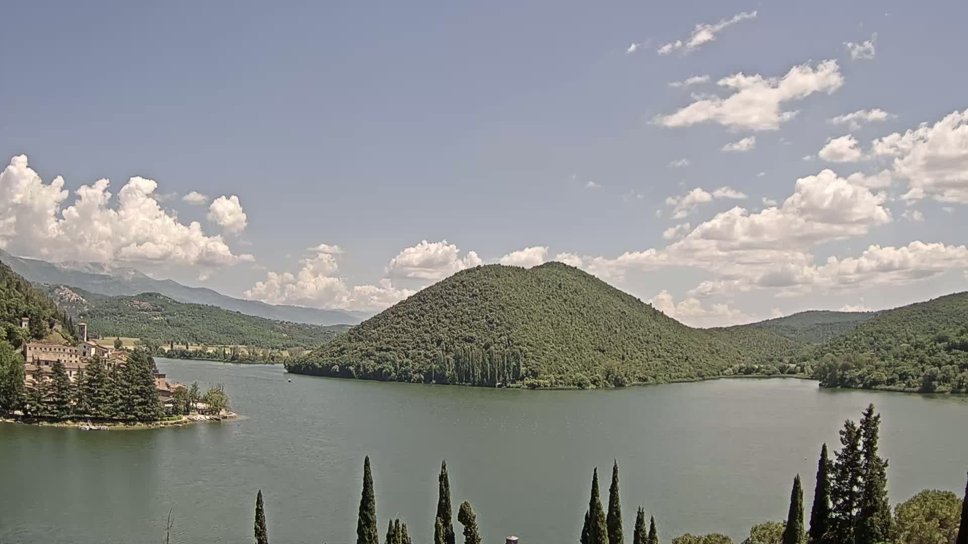 A lake surrounded by green hills and a building complex on a small island under a partly cloudy sky.